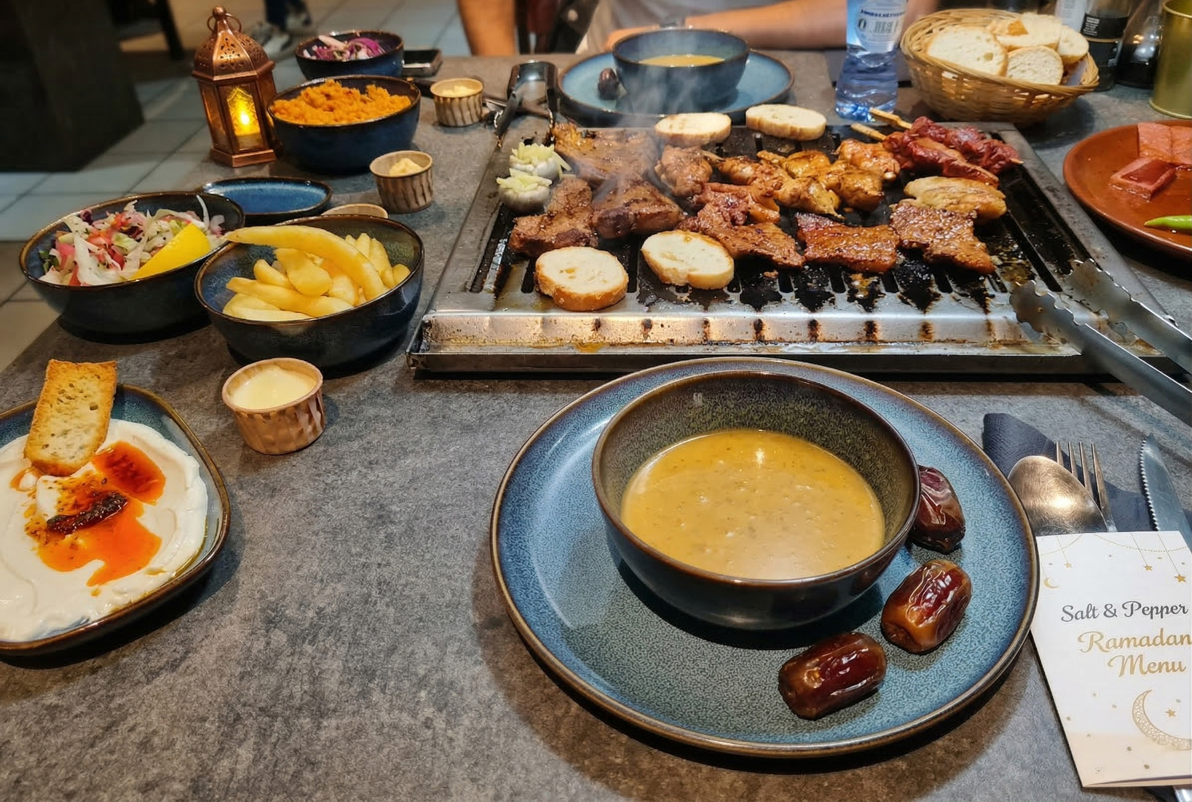 Een overvolle tafel in restaurant Salt & Pepper Eindhoven met een speciaal Ramadan Iftar-menu. In het midden staat een actieve tafelgrill met gegrild vlees en brood. Eromheen staan schalen met linzensoep, hummus met chili-olie, friet, salades, een mandje met brood en dadels. Een gast zit aan de overkant met een flesje water.
