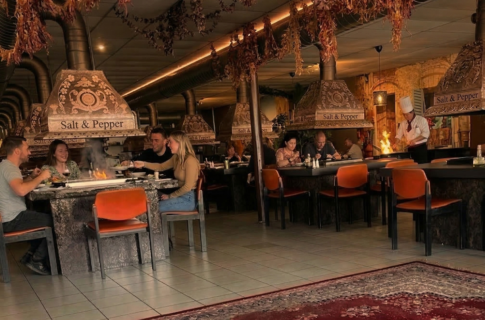 Zijaanzicht van het interieur van restaurant Salt & Pepper in Eindhoven. Meerdere grote, donkere tafels met ingebouwde grills zijn te zien, met koperkleurige afzuigkappen erboven. Een rode, geornamenteerde oosterse loper bedekt de vloer in de voorgrond.